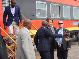 Vários homens de terno e crachá descem de um trem vermelho e amarelo enquanto cumprimentam autoridades na plataforma ferroviária.