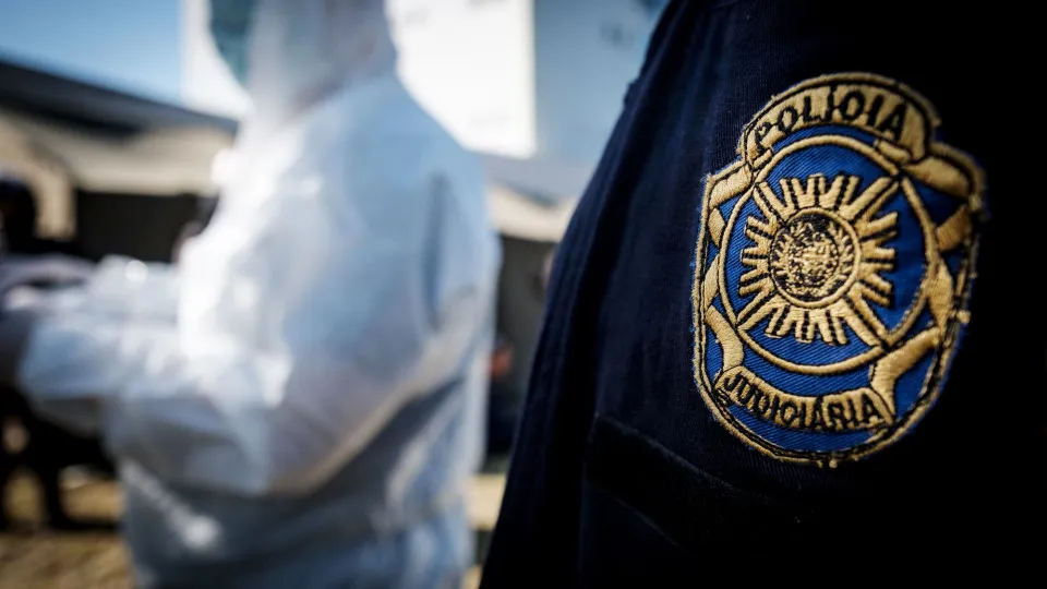 Mais próximo aparece a logo da polícia judiciária e ao fundo uma pessoa veste uma roupa branca totalmente soberto, com máscaras e luvas