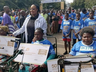 Mulheres com deficiência recebem prêmios em triciclos adaptados após participação em corrida comemorativa, cercadas por uma multidão de participantes e apoiadores usando camisetas azuis.