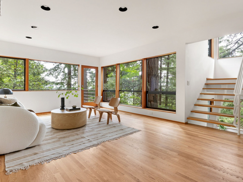 Remodeled open concept living room with white walls, stairs, wood ceilings and floor, and large windows with tree views create the feel of an open, tree house.