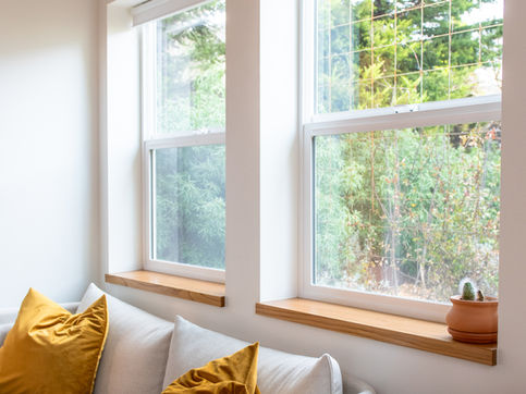 Modern double paned windows are above a soft white couch with saffron colored pillows and a small round table with chair. next to the couch.