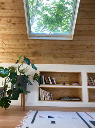 An attic renovation created a bedroom with built in shelves below a tree-filled skylight with hard wood floors and ceiling.