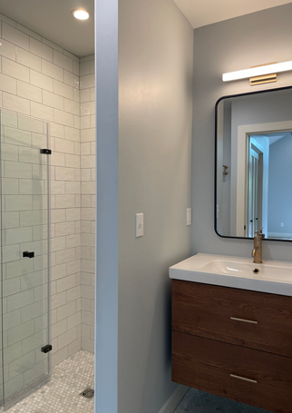 A white-tiled shower on the left is separated by a small wall from the white sink on a dark cabinet and simple metal framed rectangle mirror above.