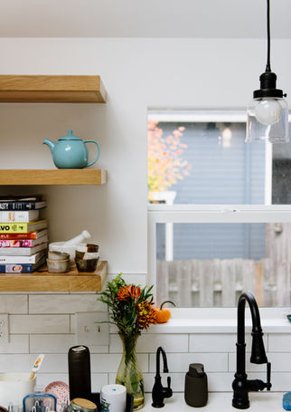 This renovated kitchen has with walls and trim around the window above the sink, dark metal faucets, and wooden floating shelves above the counter to the left.