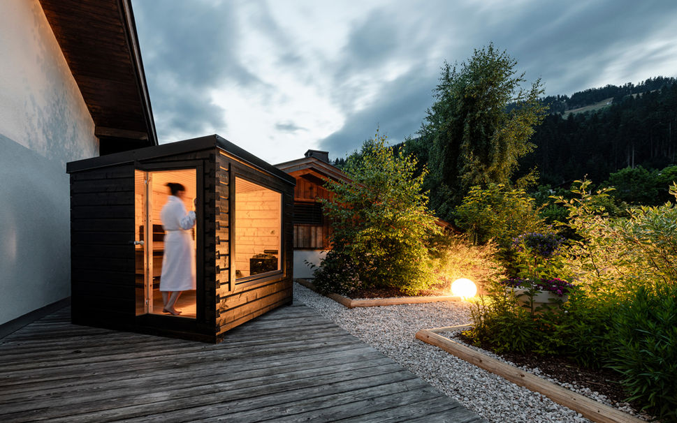 Hetki outdoor Finnish log sauna in hotel garden at dusk with lights on, person entering, evening wellness haven oasis in Finland.