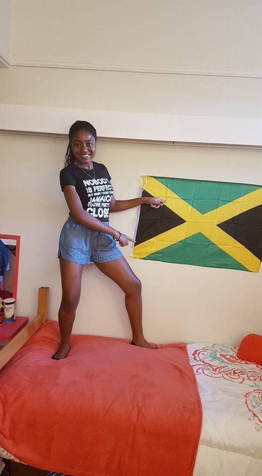 Jenine is standing on her bed in a t-shirt and shorts and is pointing to her Jamaican flag on her wall.