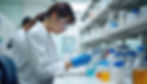 Pharmacist in goggles and gloves examines test tube in lab, surrounded by bottles on shelves. Focused and analytical mood.