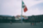 People walking near the Mexican flag that's on a pole with buildings and colorful houses in the background