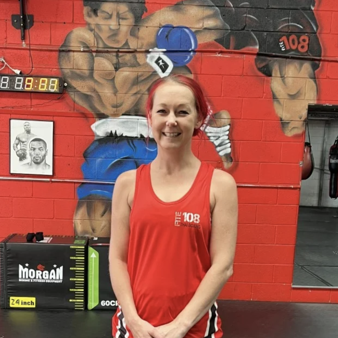 Amy Corry in a red Fite108 tank top stands in a gym with boxing mural on red brick wall.