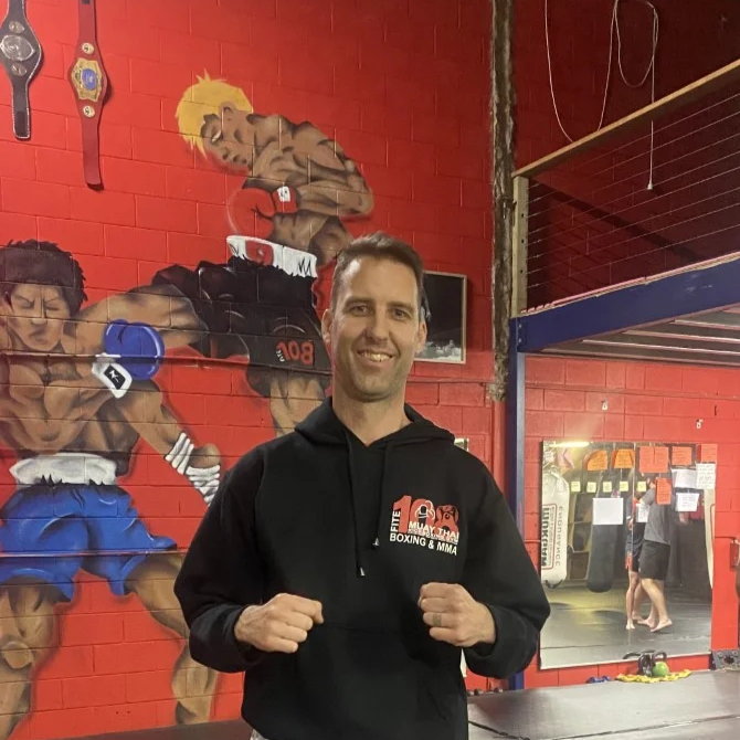 Dave Vales in Fite108 gear stands on gym mat. Mural of boxers behind.