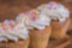 Three cupcakes with white frosting and rainbow sprinkles on a wooden surface