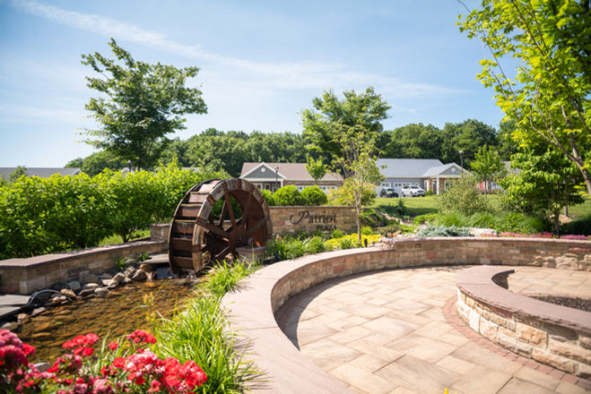 Water wheel feature with stone patio, flowers, and residential homes in background.