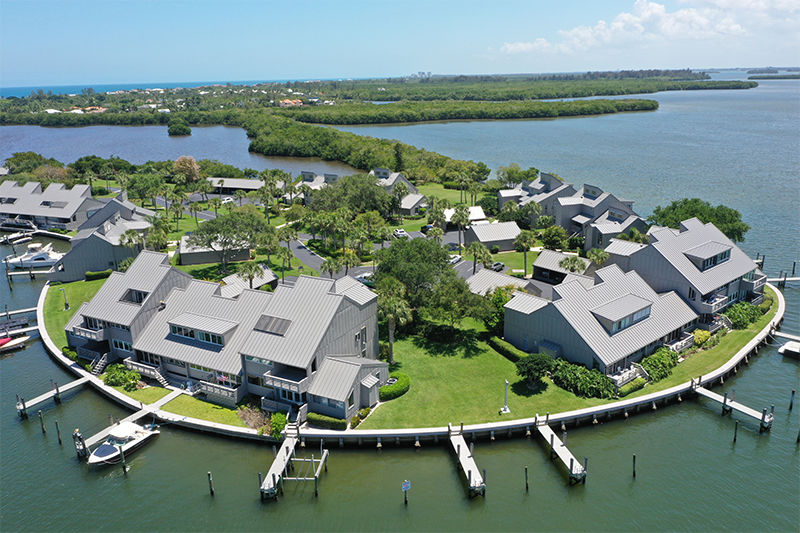 Aerial view of coastal residential buildings with light gray metal roofing, arranged along a curved waterfront with docks and surrounding waterways.