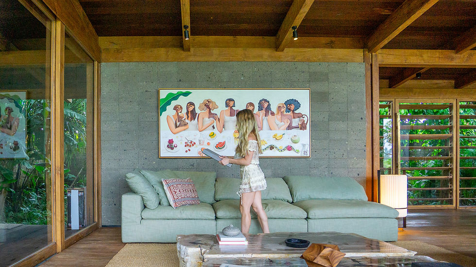 Woman in white dress stands by a pale green sofa in a wood-and-glass living room, studying colorful wall art.