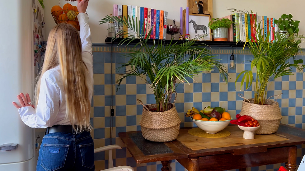 Blonde woman in kitchen reaching for hanging fruit basket. Blue-checkered tiles, shelf with books, and a table with plants and fruit.