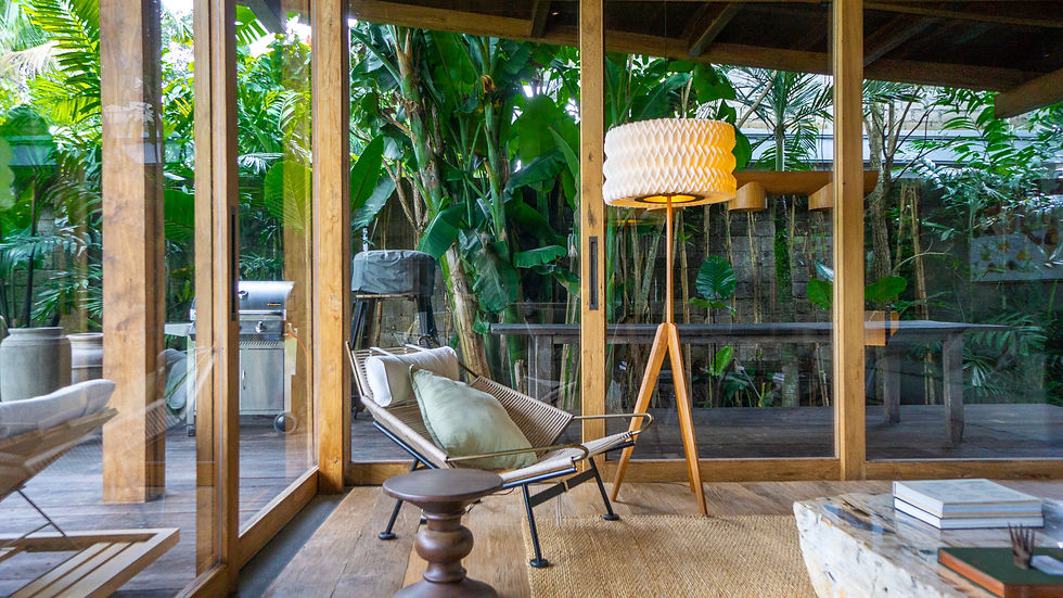 Tropical patio living room with wood-framed glass walls, lounge chair, floor lamp, and lush green plants outside.