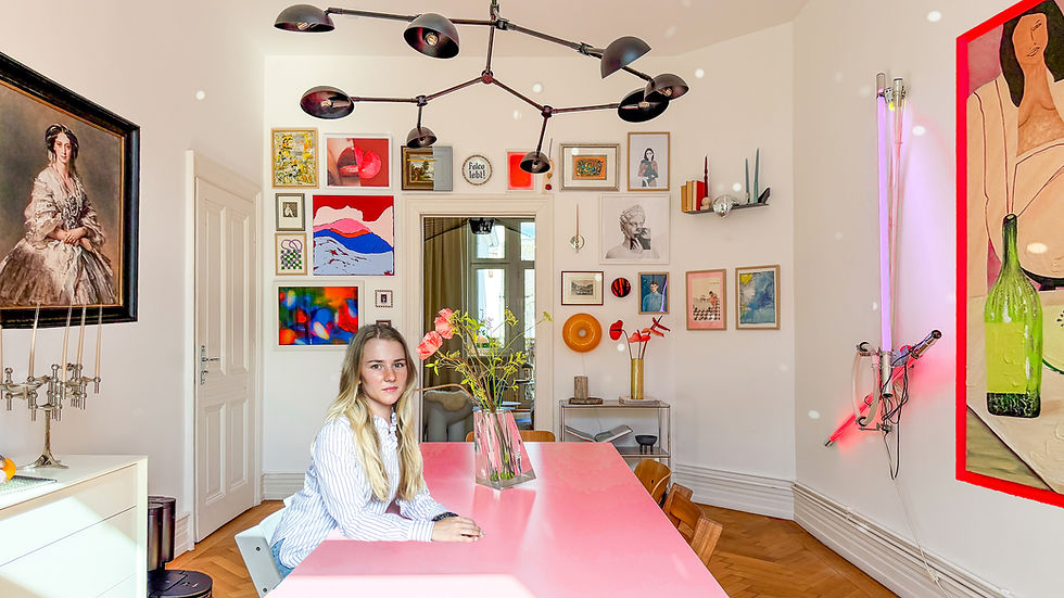 Woman sits at a pink table in a bright art-filled room with colorful framed paintings, candles, and a modern black ceiling light.