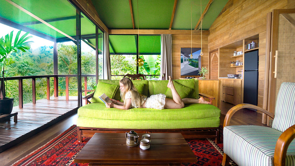 Woman reading on a lime-green sofa in a wood-paneled tropical cabin, with open windows, lush greenery, and a cozy mood.