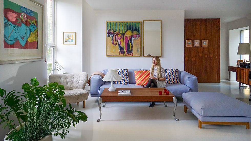 Woman sits on a lavender sofa in a bright modern living room, surrounded by colorful artwork, plants, and a wooden coffee table.