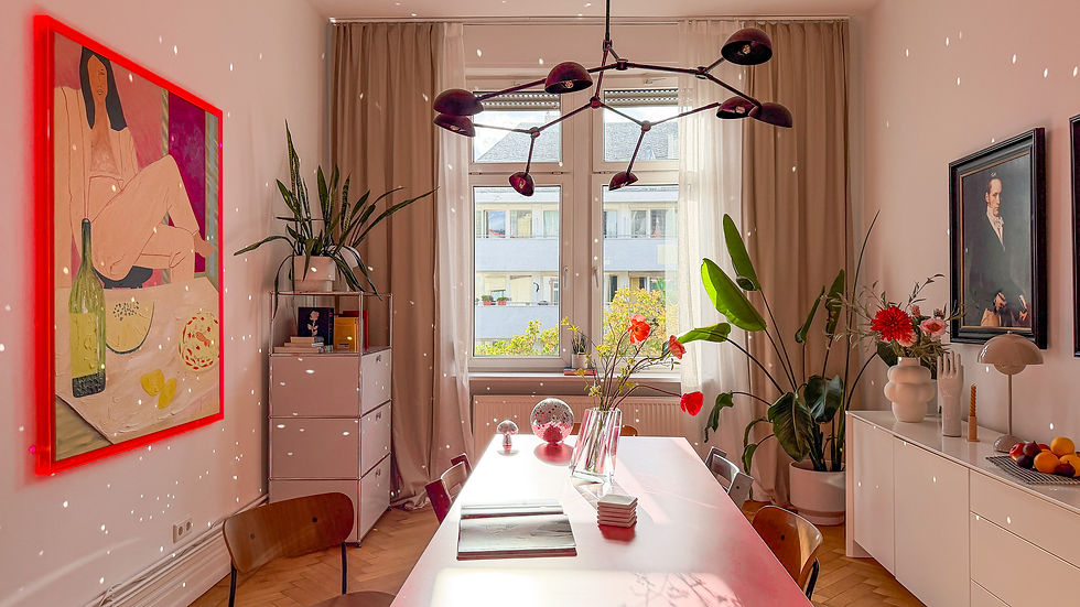 Sunlit modern dining room with red-framed art, plants, white table, sculptural lamp, and portraits by curtained windows.