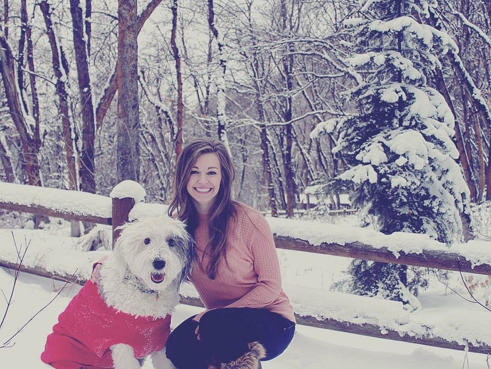 Landscape photo of a smiling woman kneeling in the snow next to a fluffy white dog in a red sweater