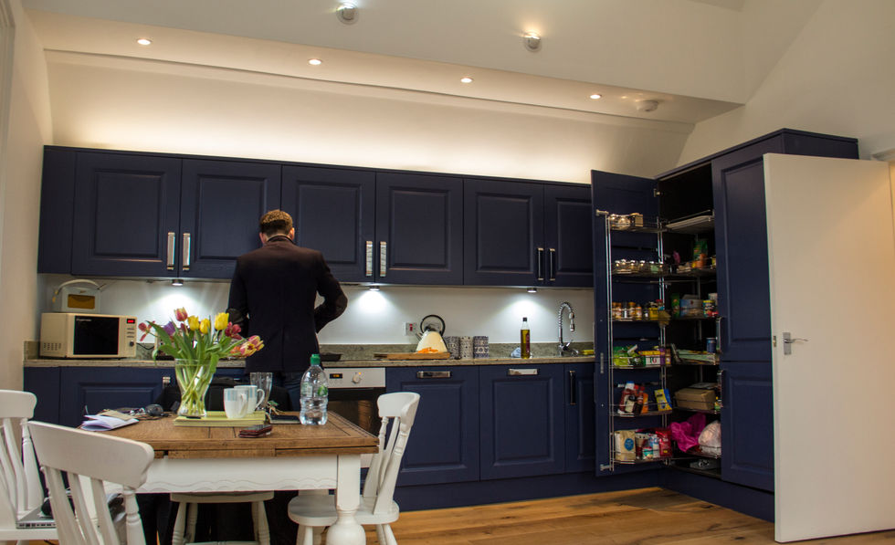 Man in a dark suit standing in a modern kitchen with dark blue cabinets, some open to reveal organized contents. A dining table and chairs are in the foreground.