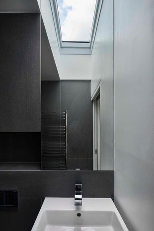 A modern bathroom with a concrete sink, a large mirror, and a skylight above. The room has a minimalist design with dark gray walls and black fixtures.