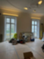 Room under renovation with a chandelier, tools, boxes, and a mirror reflecting a person. Large windows show an outdoor view.