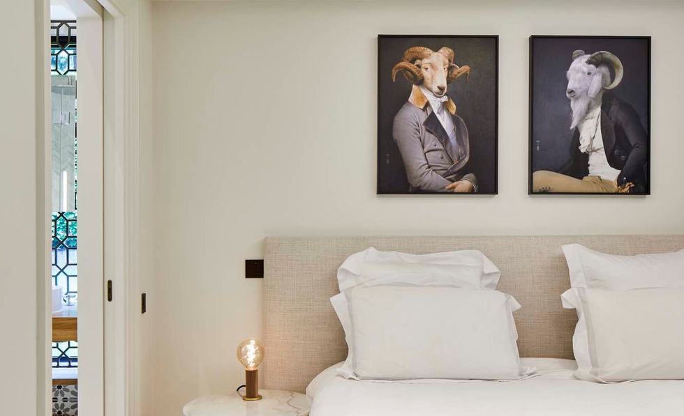 A bedroom with a double bed covered in a blue bedspread. Two framed paintings hang above the bed. A bedside table with a lamp is located next to the bed.