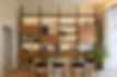 Modern dining room with a glass table, black and pink chairs, and a floor-to-ceiling shelving unit with warm lighting, wooden cabinets, and decorative plants.