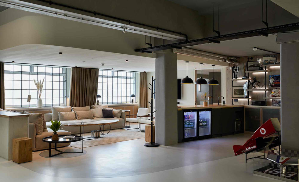 A modern living room with a comfortable sofa, a coffee table, and a small bar area with mini-fridges. The room has large windows and exposed brick walls.