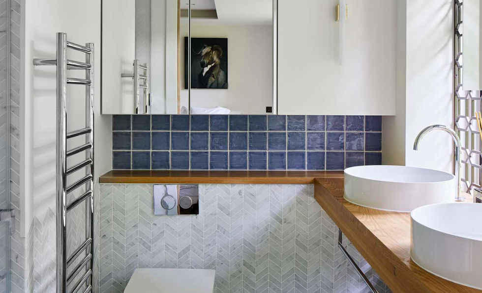 The image shows a modern bathroom with geometric patterned tiles, a wooden countertop holding two vessel sinks, and a toilet positioned beneath a mirrored cabinet. The space is bright, featuring a combination of blue, white, and natural wood tones.