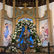 Auf dem Bild sieht man einen reich verzierten Altar in einer Kapelle/Kirche: In der Mitte steht eine Marienstatue mit Krone, die das Jesuskind auf dem Arm trägt. Die Figur ist von einem großen Kranz aus weißen Blumen umrahmt; links und rechts sind gemalte Engel/Figuren zu sehen, davor ein schmiedeeisernes Gitter.