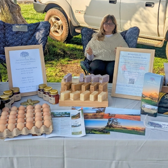 A young woman with glasses sits behind a market stall table laden with produce. On the white tablecloth are stacks of egg cartons, a tray of loose brown eggs, several jars of honey, bars of handmade soap, and framed price lists and informational signs about "Pasture-Raised Eggs" and a "Market Bundle." Pamphlets with pictures are also spread out. In the background, there is a white pickup truck parked on grass under sunlight.