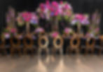 Luxury head table setup with dramatic pink, purple, and blue floral arrangements, black velvet tablecloth, and gold chairs.