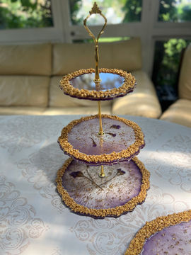 Expositor de repostería de tres niveles con platos circulares de resina en tonos púrpura y blanco, unidos por un eje central dorado y rematados con bordes texturizados en oro.