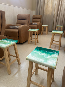 Perspectiva de una sala decorada con varios taburetes de madera, cada uno con diseños únicos de resina en tonos verdes y azules inspirados en el océano.