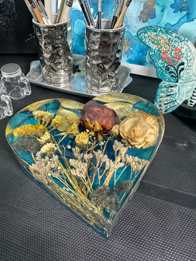 Vista en ángulo de un corazón de resina donde se aprecian flores preservadas, incluyendo una pequeña rosa y follaje silvestre, creando un efecto tridimensional de jardín eterno.