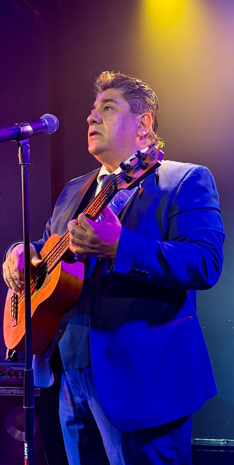 Male vocalist and guitarist performing live on stage with a Mexican trio under colorful concert lighting.