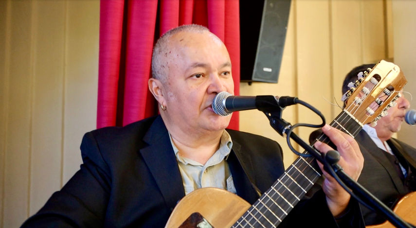 Lead vocalist of a Latin trio singing into a microphone while playing acoustic guitar at an event.