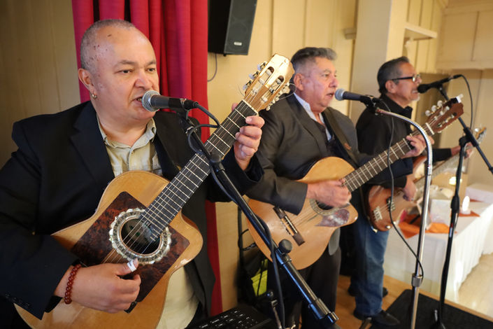 Latin trio musicians performing live with acoustic guitars and microphones at an indoor event.