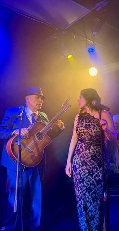 Male vocalist and female singer performing live with a Mexican trio guitarist on stage under blue and yellow concert lights.