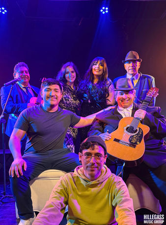 Group of musicians and performers posing together on a stage with colorful lighting during a live Latin music performance.