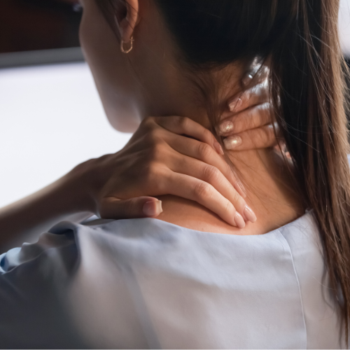 Woman in light blue shirt massaging her neck with both hands, appearing tense. Soft lighting, blurred background, indoor setting.