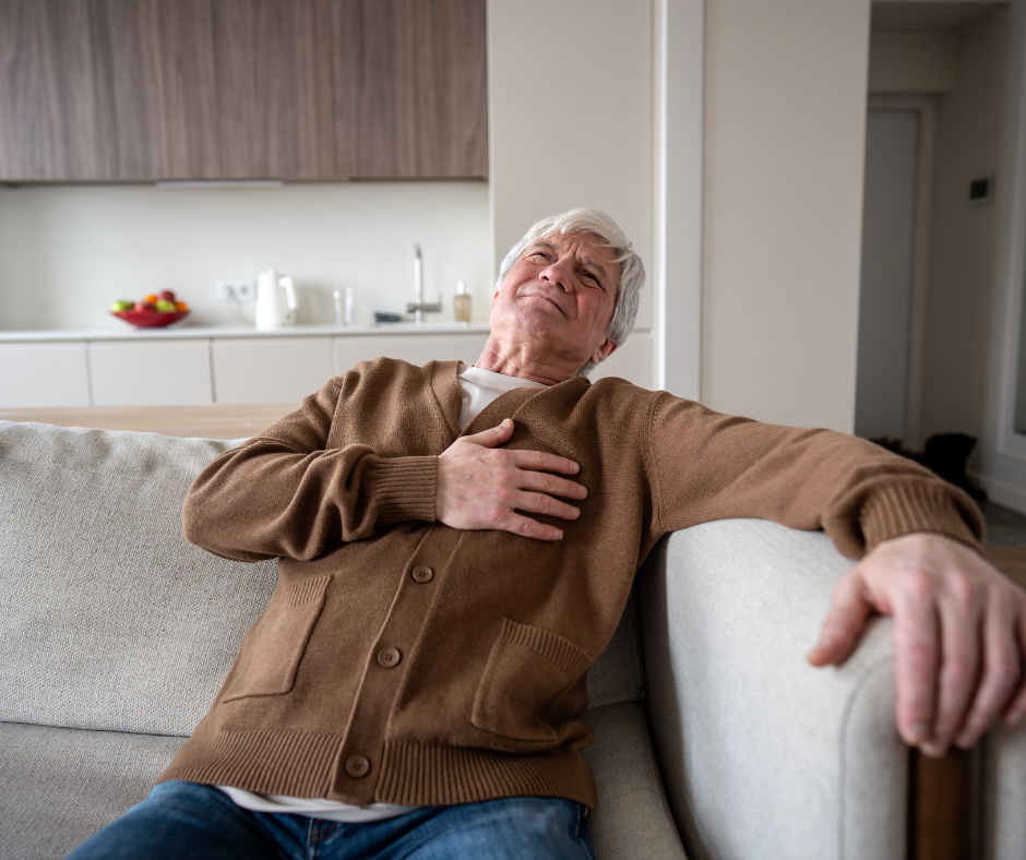 Elderly man clutching chest in pain on a couch in a modern kitchen. Wearing brown cardigan and jeans, expression of discomfort.