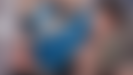 Man in a gym, wearing a blue tank top and black shorts, clutching his lower back in pain, near a kettlebell. Background blurred.