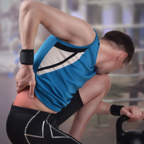 Man in a gym, wearing a blue tank top and black shorts, clutching his lower back in pain, near a kettlebell. Background blurred.