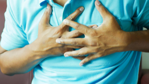 Man in a light blue shirt clutches chest in discomfort, sitting on a maroon couch. The background is plain, suggesting an indoor setting.