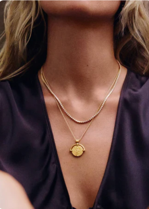 Close-up of a woman in a dark V-neck top wearing layered gold necklaces, one with a round pendant.