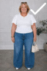 Smiling woman in white top and wide-leg blue jeans holds a beige handbag in a gray room with a potted plant.